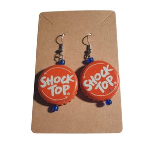 May include: A pair of handmade earrings featuring orange bottle caps with the text "Shock Top" in white. The earrings have silver-colored hooks and small blue beads. The earrings are displayed on a brown card.