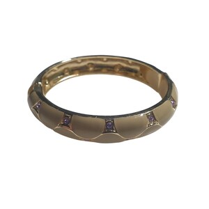 May include: Gold-toned hinged bangle bracelet with a cream-colored enamel finish. The bracelet features a repeating pattern of scalloped sections, each accented with a small, square-set purple gemstone. A classic jewelry piece.