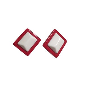 May include: A pair of diamond-shaped earrings. Each earring features a white, rectangular center set within a red, diamond-shaped frame. The earrings are set against a white background.