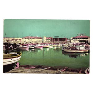 May include: Vintage postcard showing a harbour scene with many boats moored in the water. The boats are painted in various colours, including pink and white. Buildings line the background under a teal sky. A wooden dock is in the foreground.