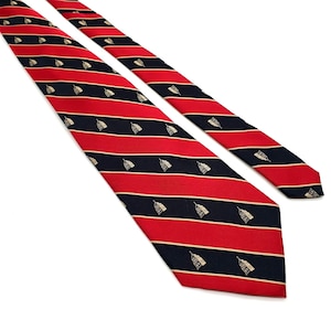 Puede incluir: Una corbata a rayas rojas y azul marino con un patrón repetido de pequeñas ilustraciones de campanas beige. La corbata está en ángulo, mostrando toda la longitud y el diseño. Las rayas están espaciadas uniformemente.