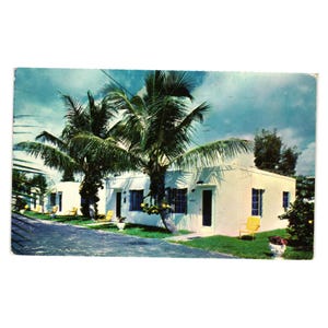 May include: A white building with a yellow door and yellow chairs on the porch. Palm trees are in front of the building. The sky is blue with white clouds.