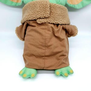 Puede incluir: Un peluche de un personaje de una popular serie de ciencia ficci&oacute;n. El juguete presenta grandes orejas verdes, un abrigo marr&oacute;n con un cuello esponjoso y pies verdes con detalles amarillos. El personaje est&aacute; sentado.