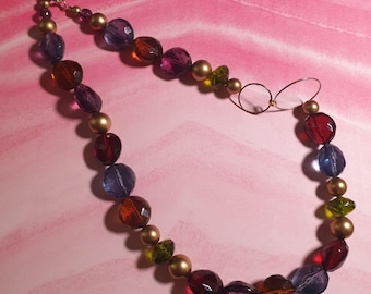 Puede incluir: Un collar colorido con cuentas facetadas en tonos rojos, morados, azules y ámbar, intercaladas con cuentas doradas. El collar tiene un delicado cierre de alambre y un pequeño colgante con marco de alambre.