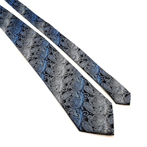 Op de afbeelding: Een blauwe en zilveren stropdas met paisley-patroon. De stropdas heeft een herhalend ontwerp van wervelende paisley-vormen in tinten blauw en zilver tegen een zwarte achtergrond. De stropdas is schuin geplaatst op een witte achtergrond.