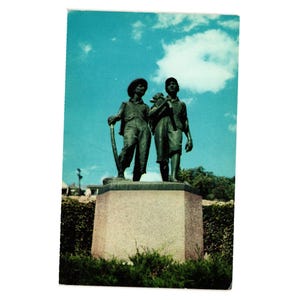 Può includere: Cartolina vintage con una statua in bronzo di due giovani figure, una con un attrezzo e l'altra che trasporta un animale. La statua è su una base di pietra contro un cielo blu con nuvole.