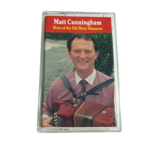 May include: A cassette tape featuring a cover with a photograph of a person playing a red accordion. The text at the top reads Matt Cunningham and West of the Old River Shannon. The person wears a light pink button-down shirt with a white collar.
