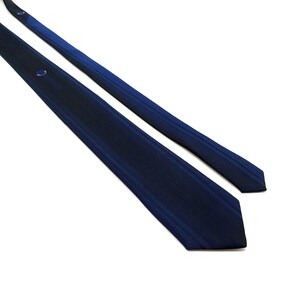 Puede incluir: Una corbata azul marino con un patrón de rayas. La corbata está en ángulo sobre un fondo blanco, mostrando su diseño. La corbata tiene un pequeño detalle bordado cerca del nudo.