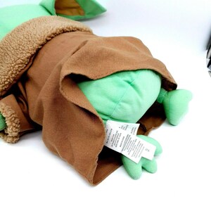 Puede incluir: Un peluche de un personaje de una popular franquicia de ciencia ficci&oacute;n. El juguete es de color verde claro con orejas grandes, y lleva un abrigo marr&oacute;n con un forro beige y difuso. La etiqueta del juguete es visible.