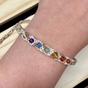 May include: A white macrame bracelet with seven colourful beads. The beads are purple, blue, green, yellow, orange, and red.