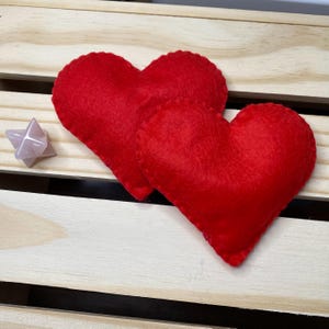May include: Two red felt hearts on a wooden surface. The hearts are sewn together and have a slightly textured surface.