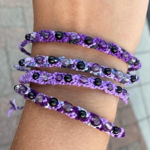Puede incluir: Cuatro pulseras superpuestas con cuentas moradas y negras. Las pulseras están hechas de cuerda y tienen un patrón tejido.