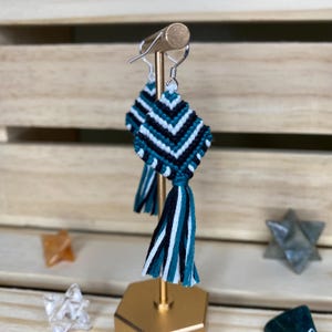 May include: Teal, black, and white geometric patterned earrings with a tassel. The earrings are displayed on a gold stand.