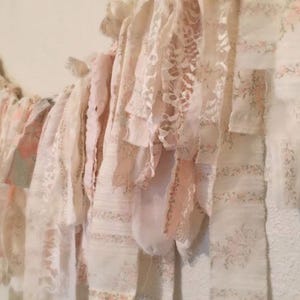 May include: A garland of layered fabric strips in shades of white and pale pink. The strips feature lace, floral patterns, and ruffled edges, creating a delicate, vintage aesthetic. The garland is hanging against a neutral wall.