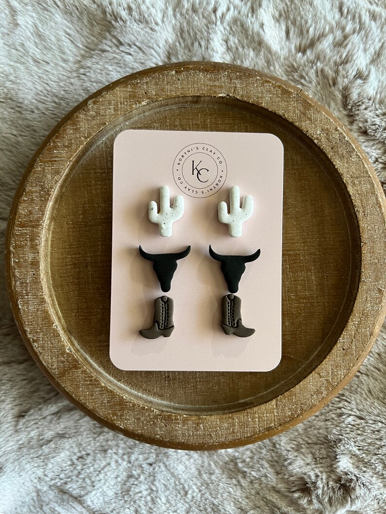 May include: A set of three pairs of stud earrings on a light pink display card with the text Kortni's Clay Co. The earrings feature white speckled cactus shapes, black bull skull silhouettes, and brown cowboy boot designs with detailed stitching.