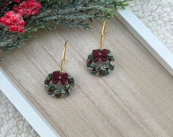 May include: A pair of dangle earrings featuring circular wreath charms. Each wreath is crafted with dark green leaves, small red berries, and gold glitter accents, topped with a dark red bow and a small clear rhinestone center on a gold hoop.