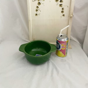May include: A green ceramic bowl with handles and a can of pink and yellow flavored sparkling water with a straw. Behind them is a cream-colored cabinet with a vine and leaf design.