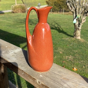 May include: A terracotta-colored ceramic pitcher with a slender, curved handle and a spout. The pitcher has a smooth, glossy finish and sits on a wooden railing. The background features a grassy area and trees.