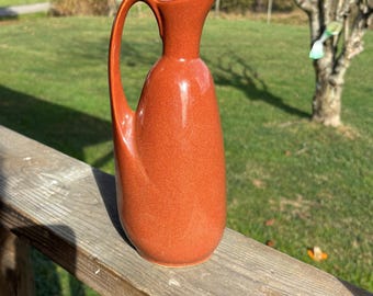 May include: A terracotta-colored ceramic pitcher with a slender, curved handle and a spout. The pitcher has a smooth, glossy finish and sits on a wooden railing. The background features a grassy area and trees.