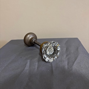 May include: A door knob featuring a clear, faceted glass handle with a starburst pattern in the center. The handle is attached to a metallic, bronze-toned circular base and a matching bronze-toned spindle extending from the back of the assembly.