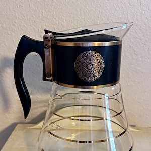 May include: A vintage glass coffee carafe with a black and gold design. The carafe has a black handle and lid, with gold accents and a decorative gold pattern on the front. Gold horizontal stripes adorn the clear glass body.