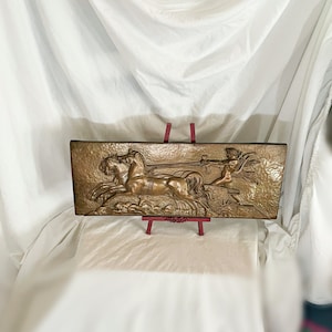May include: Bronze rectangular relief sculpture depicting a chariot pulled by two horses, with a winged figure holding reins. The artwork is displayed on a red easel against a white backdrop.