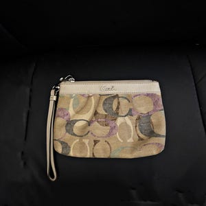 May include: A beige Coach wristlet featuring a multi-colored signature C pattern in purple, blue, and brown. The pouch has a top zipper closure, a light beige leather trim, and a matching leather wrist strap attached to the side hardware.