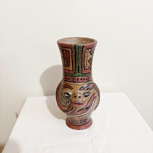May include: A hand-painted ceramic vase with a face design. The vase features a brown base, a tan midsection with a face, and a red and green patterned top. The face has red lips and black details.