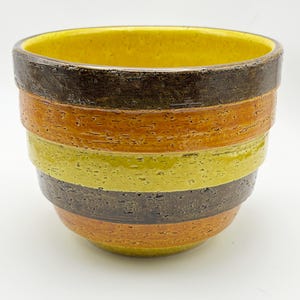 May include: A ceramic planter with a textured, banded design. The interior is yellow, with bands of brown, orange, and green on the exterior. The planter has a wide opening and a slightly tapered shape, suitable for plants.