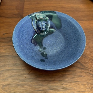 May include: A small, round, blue ceramic bowl with a face sculpted on the side. The bowl has a textured surface and a glossy finish. The face is a darker shade of blue, adding depth to the design. The bowl is sitting on a wooden surface.