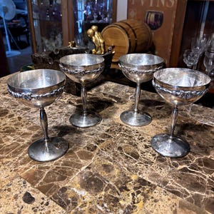 May include: A set of four silver-toned metal goblets with a wide, shallow bowl and a slender stem. Each goblet features a decorative band of embossed grape and vine motifs encircling the exterior rim of the bowl, resting on a circular base.