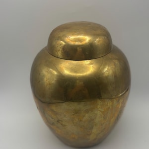 May include: A golden-colored, round jar with a matching lid. The jar has a bulbous body and a slightly textured surface. The lid sits snugly on top, creating a cohesive, antique aesthetic. The jar is likely made of metal.