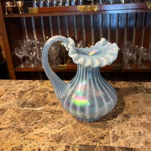 May include: A light blue, iridescent glass pitcher with a ruffled rim and a curved handle. The pitcher has vertical ridges and sits on a brown marble surface. The pitcher is a decorative item.