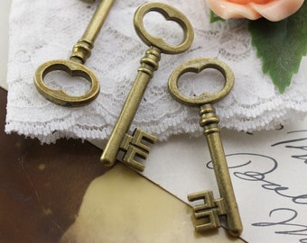 May include: Three antique-style bronze keys with ornate designs. The keys have different head shapes, including oval and keyhole designs. The keys are arranged on a white lace surface, with a peach-colored rose and green leaves in the background.
