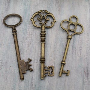 Könnte beinhalten: Drei Schlüssel im antiken Stil aus bronzefarbenem Metall. Die Schlüssel weisen verschiedene verzierte Designs auf, darunter dekorative Schlüsselköpfe und -schäfte. Sie liegen auf einer hellblauen und weißen Holzoberfläche und erinnern an eine Vintage-Ästhetik.