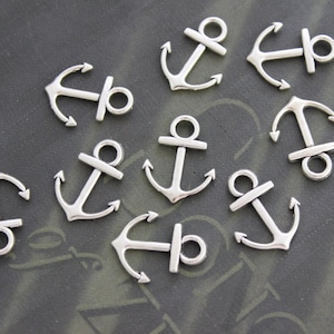 Könnte beinhalten: Eine Sammlung von silberfarbenen Anker-Anhängern aus Metall. Die Anhänger sind klein und detailliert, mit einem klassischen Anker-Design.