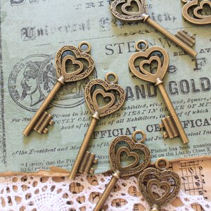 May include: A collection of antique-style key charms with ornate heart-shaped designs. The keys are made of metal and have a distressed, aged finish. The keys are arranged on a vintage-looking paper background with text.