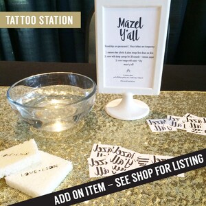 May include: A white sign with the text "Mazel Y'all" and instructions for applying temporary tattoos. The sign is on a gold glitter table with a bowl of water and several temporary tattoos. The text "ADD ON ITEM - SEE SHOP FOR LISTING" is at the bottom of the image.