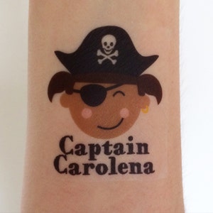 May include: A temporary tattoo of a cartoon pirate girl wearing a black pirate hat with a skull and crossbones design. The girl has brown hair, a black eye patch, and a gold earring. The text "Captain Carolena" is printed below the image.