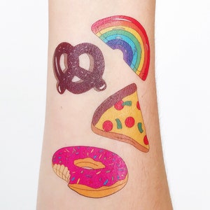 May include: Four temporary tattoos on a person's arm. The tattoos are of a rainbow, a pretzel, a slice of pizza, and a donut. The donut is pink with sprinkles and has a bite taken out of it.