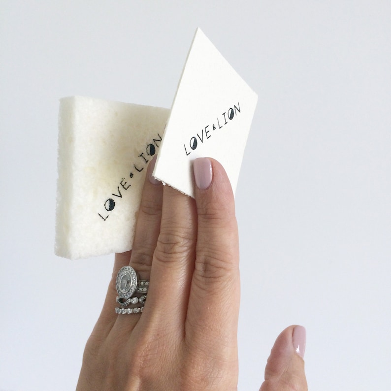 May include: Two white cleaning cloths with the text "LOVE & LION" printed on them. A hand is holding the cloths.