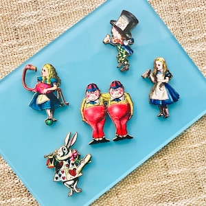 May include: A set of six Alice in Wonderland themed wooden pins. The pins feature characters from the book, including the Mad Hatter, the Queen of Hearts, the Cheshire Cat, and the White Rabbit. The pins are painted in bright colours and have a whimsical design.