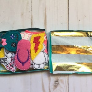 May include: A small, open teal box filled with colorful felt appliques. The appliques include a pink flip-flop, a yellow lightning bolt, and a teal bow. The box is lined with gold and white striped tissue paper.
