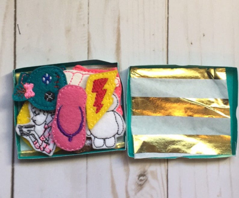 May include: A small, open teal box filled with colorful felt appliques. The appliques include a pink flip-flop, a yellow lightning bolt, and a teal bow. The box is lined with gold and white striped tissue paper.