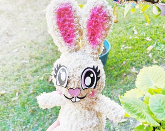 May include: A plush bunny toy with cream-colored fur and pink accents on the ears, cheeks, and nose. The bunny has large, expressive eyes with long eyelashes and a small, smiling mouth. The toy is held in a hand against a background of green grass and foliage.