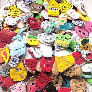 May include: A collection of colorful felt appliques in various shapes and designs. The appliques include cartoon characters, food items, and other whimsical shapes. Some appliques have embroidered details and text, such as "SOAR" and "Tiffany & Co."