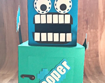 May include: A blue and green cardboard robot box with a white crown, large eyes, and a toothy grin. The box has a hinged lid and a label that says "Cooper".
