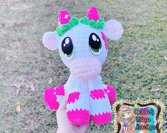 May include: A handmade white and pink crochet cow plushie with large black and green eyes. The cow has pink spots, ears, and horns with a green leafy crown. The plushie is held in a hand, with a Kendra's Unique Boutique logo in the background.