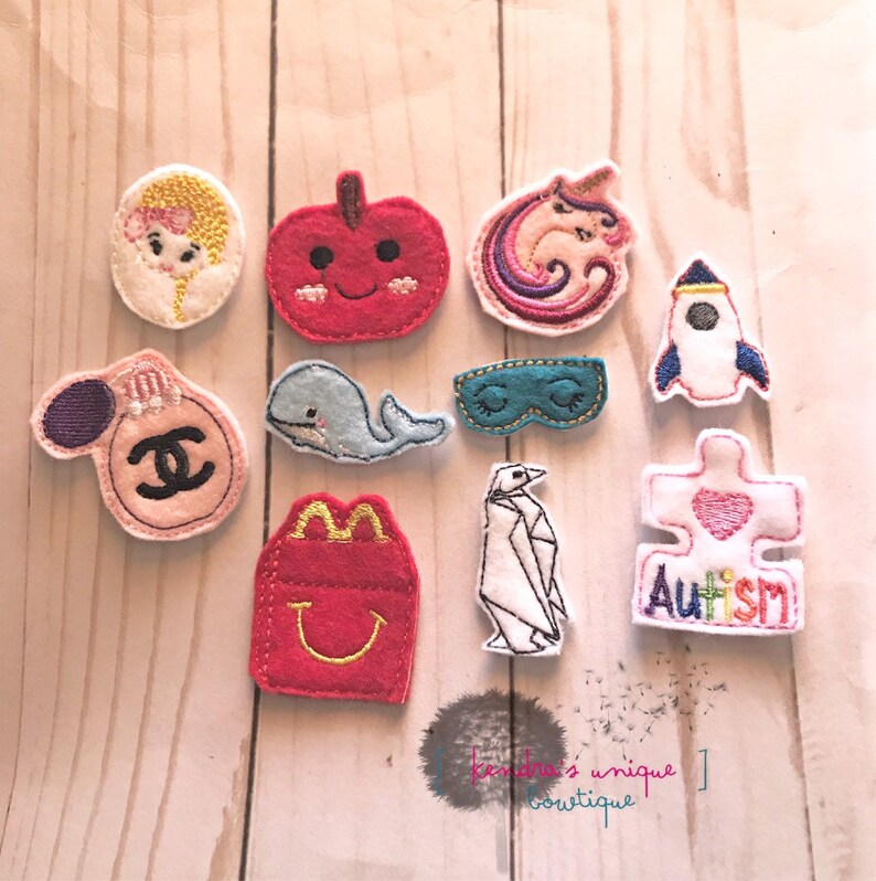 May include: A collection of colorful felt appliques in various shapes, including a girl, apple, unicorn, rocket, perfume bottle, whale, sleep mask, fries, penguin, and puzzle piece with the word "Autism".