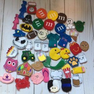 May include: A collection of colorful felt appliques in various shapes and designs. Includes characters like Darth Vader, Batman, and Pac-Man, along with food items, a tractor, and other novelty shapes. Perfect for crafting or embellishments.
